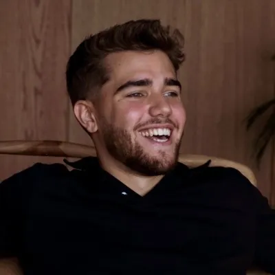 Smiling young man with short brown hair and beard sitting on a chair indoors against a wooden background.