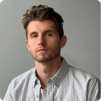 Young man with short brown hair and a light beard wearing a striped button-up shirt against a plain gray background.